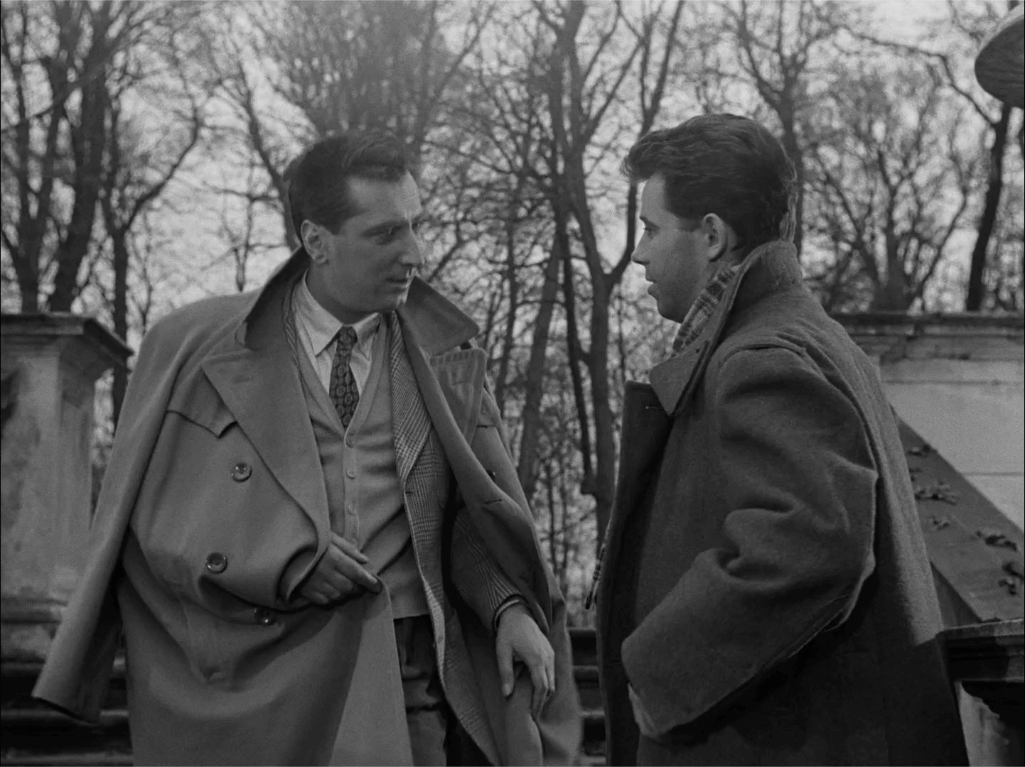 Dwóch mężczyzn w płaszczach stoi na zewnątrz przed kamiennymi schodami i nagimi drzewami, pogrążonych w poważnej rozmowie. Jeden z mężczyzn zarzuca płaszcz na ramię, a drugi stoi naprzeciwko niego, obaj wydają się pogrążeni w rozmowie.