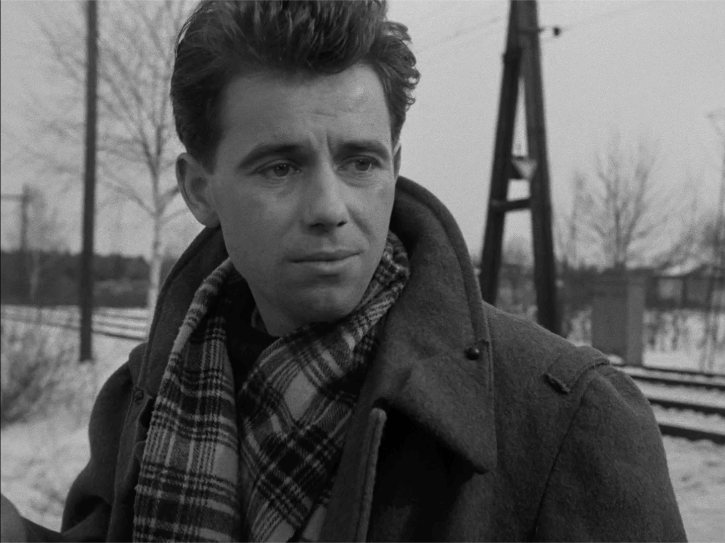 Młody mężczyzna o ciemnych włosach, ubrany w płaszcz i kratę, stoi na zewnątrz, w pobliżu pokrytych śniegiem torów kolejowych w zimny, pochmurny dzień. W tle widać nagie drzewa i słupy energetyczne.