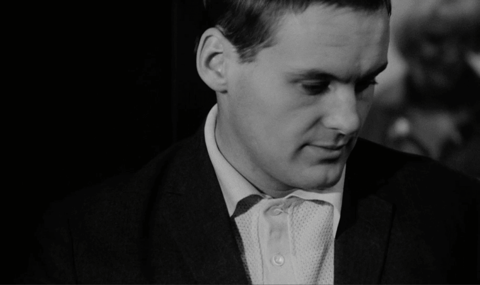 Mężczyzna w garniturze i wzorzystej koszuli patrzy w dół z poważnym wyrazem twarzy. Tło jest ciemne, z niewyraźnymi kształtami i inną rozmazaną postacią widoczną. Obraz jest czarno-biały.