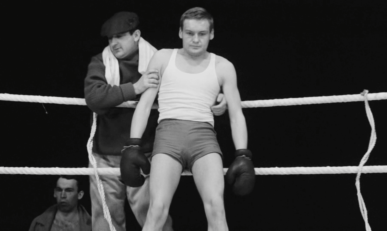 Młody bokser w rękawiczkach i spodenkach opiera się o liny ringu bokserskiego, podtrzymywany przez mężczyznę w czapce i szaliku. Inny mężczyzna stoi w tle, częściowo widoczny. Obraz jest czarno-biały.