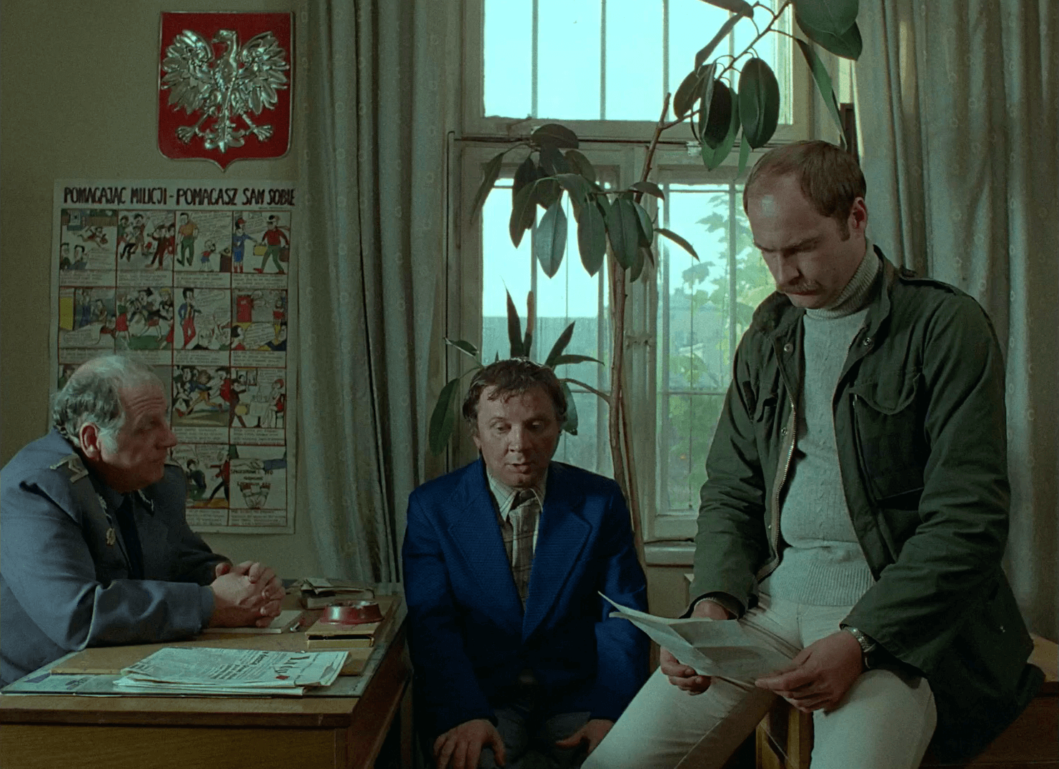 Trzech mężczyzn jest w biurze: jeden w mundurze policyjnym siedzi przy biurku, drugi w niebieskim garniturze siedzi obok niego, a trzeci mężczyzna w swobodnym ubraniu stoi i czyta dokument. W tle widać dużą roślinę i okno.