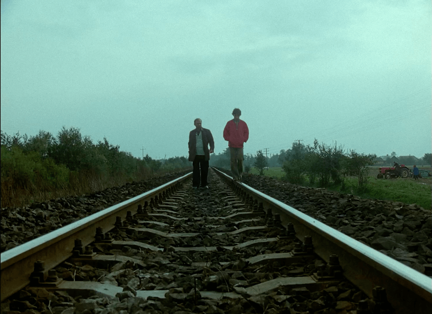 Dwie osoby idą obok siebie środkiem torów kolejowych w pochmurny dzień, w otoczeniu zielonych krzewów i drzew, w oddali po prawej stronie widoczny jest czerwony traktor.