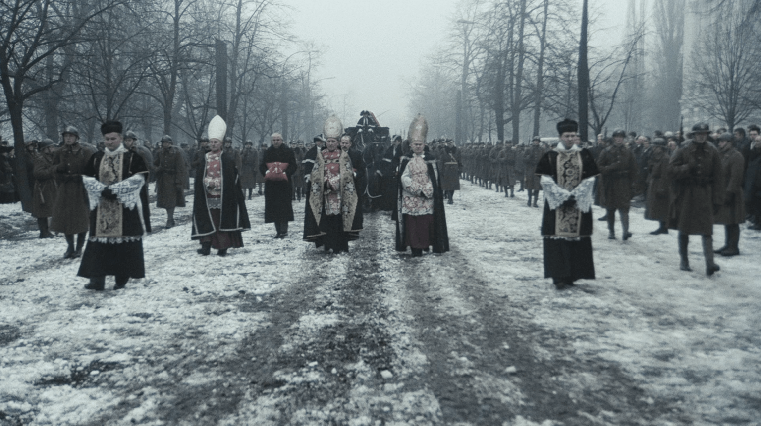 Grupa duchownych w ozdobnych szatach prowadzi kondukt pogrzebowy przez ośnieżoną, obsadzoną drzewami aleję. Za nimi idą żołnierze i żałobnicy, w tle widoczny jest karawan ciągnięty przez konie.