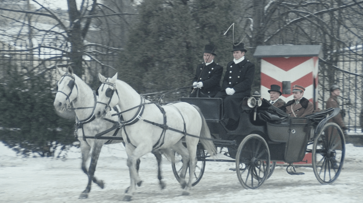 Powóz konny zaprzężony w dwa białe konie przewozi pięć osób, trzy w czarnych mundurach i dwie w brązowych. Jedzie on zaśnieżoną ścieżką wśród drzew i budki wartowniczej w czerwono-białe pasy.