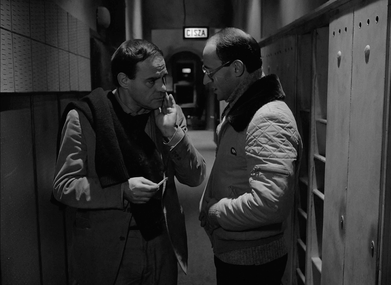 Dwóch mężczyzn stoi w słabo oświetlonym korytarzu, pogrążonych w poważnej rozmowie. Jeden z nich gestykuluje ręką w pobliżu twarzy drugiego, podczas gdy obaj wydają się skupieni. Znak nad nimi głosi CISZA (cisza) po polsku.