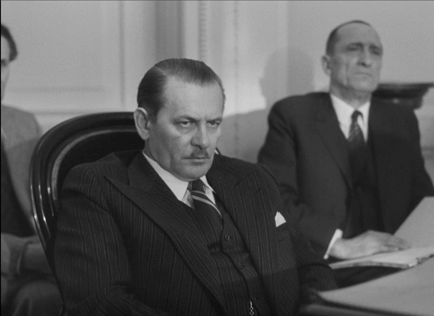 Surowo wyglądający mężczyzna w garniturze w prążki siedzi w sali sądowej, a za nim siedzi inny mężczyzna w garniturze, trzymając papiery. Obaj wydają się poważni i skupieni.