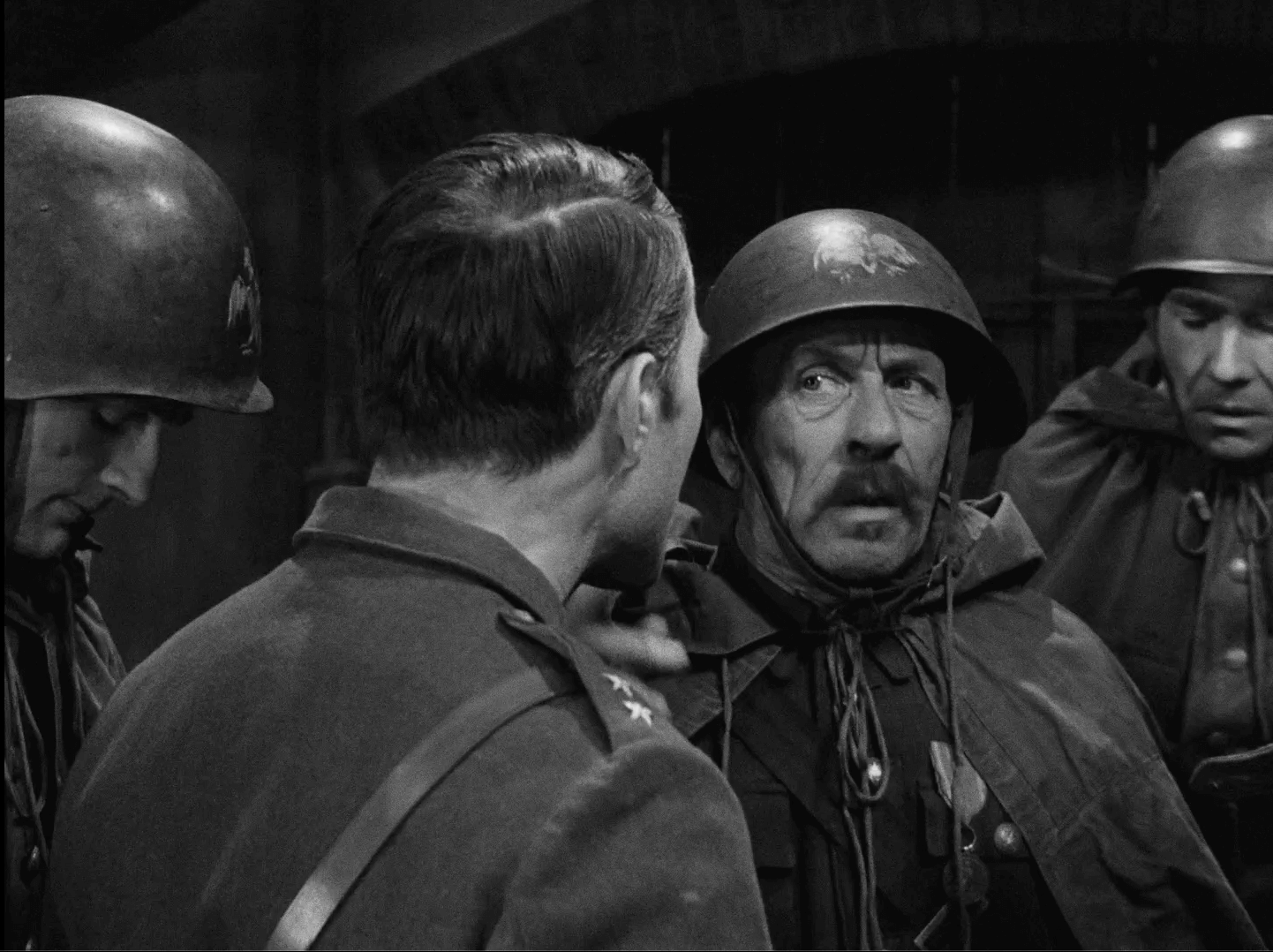 Czterech żołnierzy w mundurach wojskowych i hełmach stoi razem w pomieszczeniu. Jeden zwrócony jest twarzą w bok, drugi z wąsami wygląda na zaskoczonego lub zaniepokojonego, a pozostali patrzą w dół, co sugeruje napiętą lub poważną rozmowę.