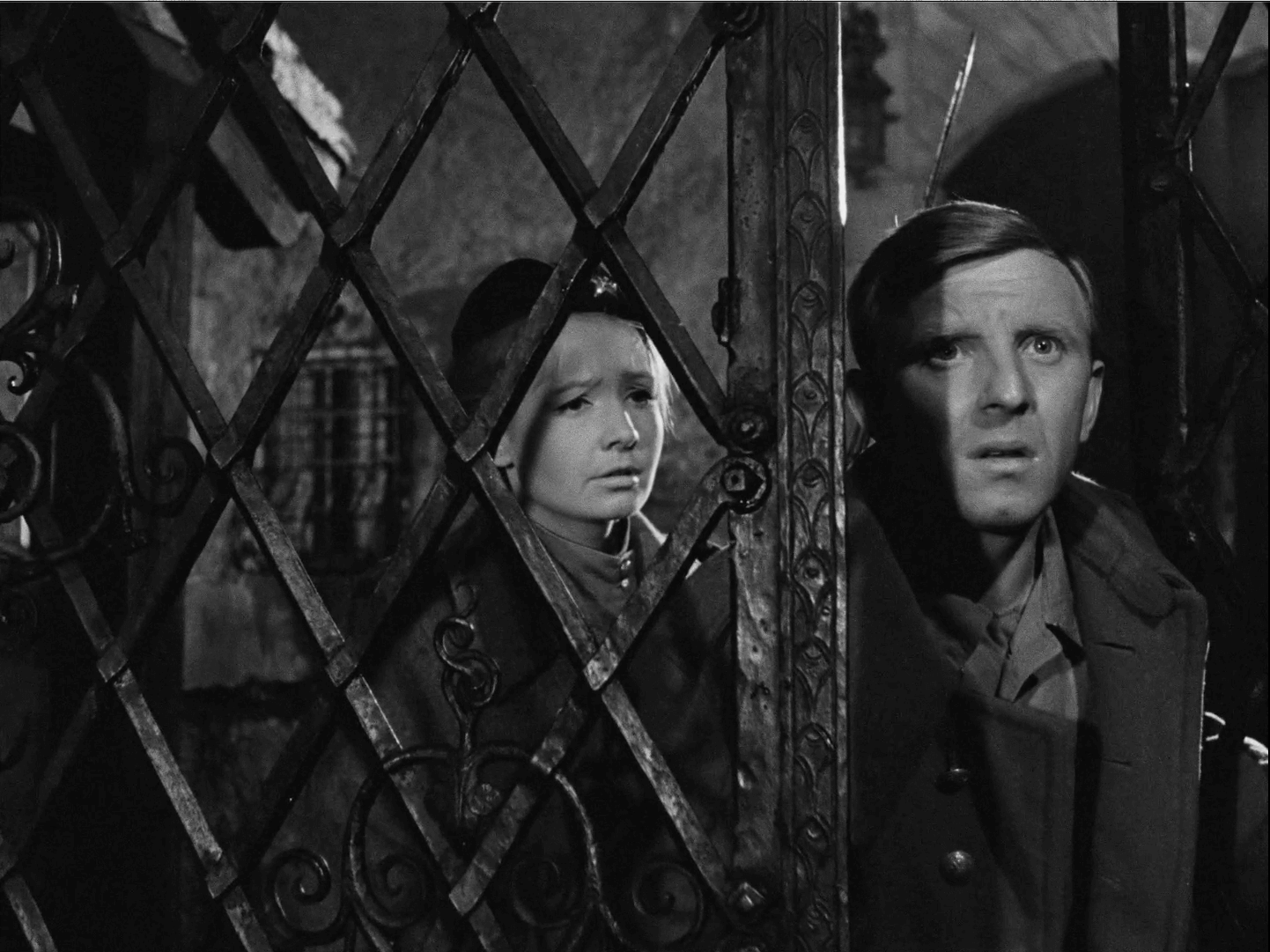Mężczyzna i kobieta stoją za dekoracyjną metalową bramą, patrząc z niepokojem w dal. Obraz jest czarno-biały, z dramatycznymi cieniami podkreślającymi ich zaniepokojone miny.