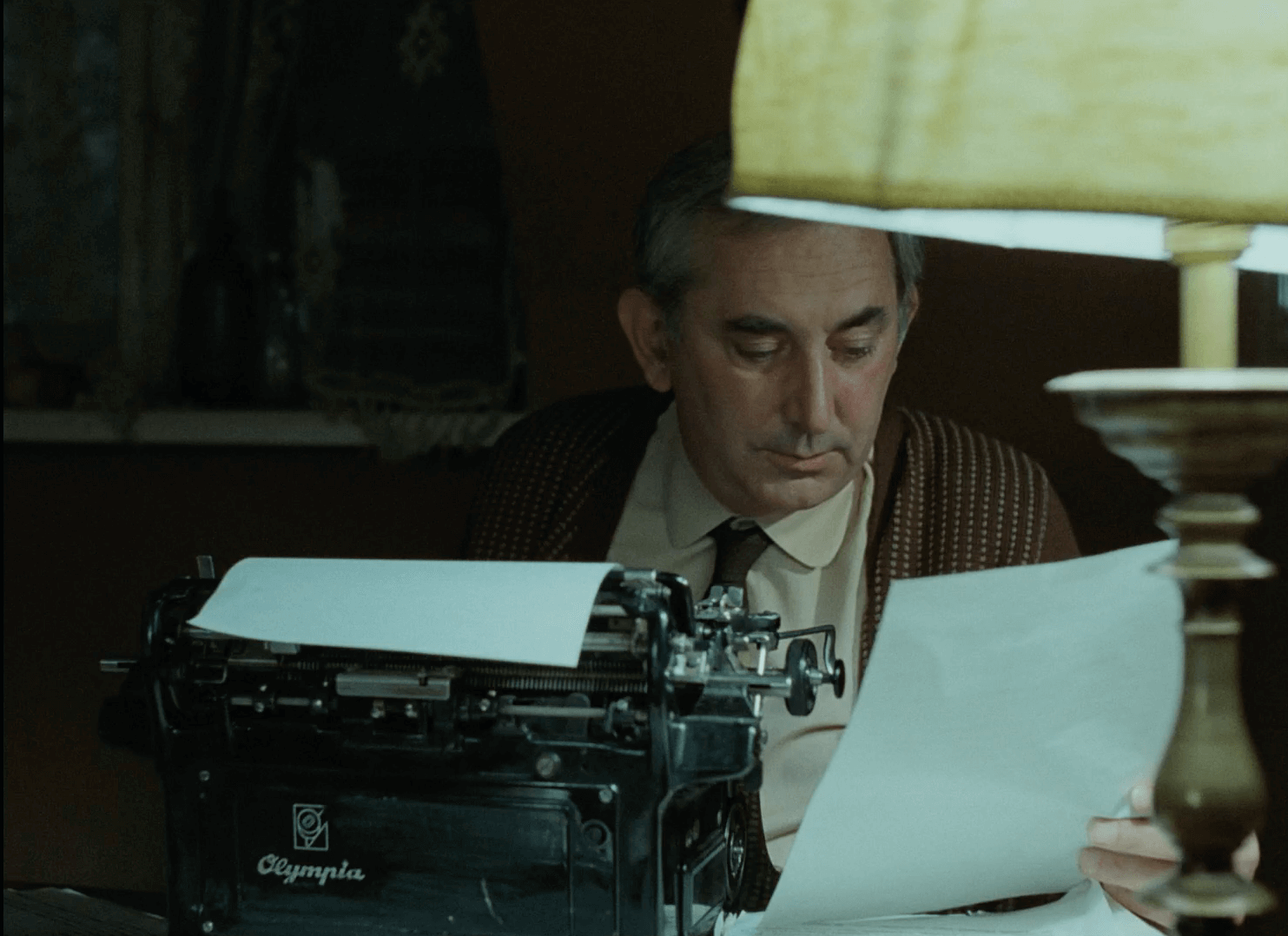 Mężczyzna siedzi przy biurku, czytając kartkę papieru obok zapalonej lampy stołowej, z maszyną do pisania Olympia przed sobą. Scena wydaje się słabo oświetlona i skupiona na jego koncentracji.