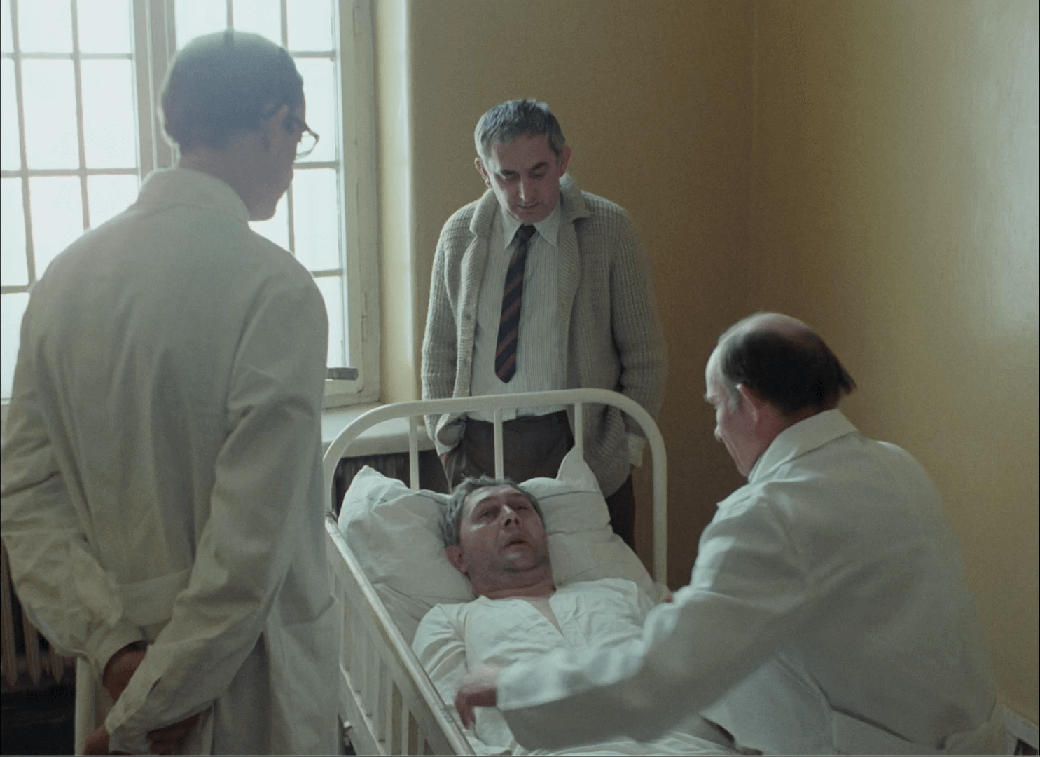 Mężczyzna leży na szpitalnym łóżku, a dwóch lekarzy w białych kitlach stoi obok; inny mężczyzna w swetrze i krawacie stoi u stóp łóżka. Promienie słoneczne wpadają przez duże okna w tle.