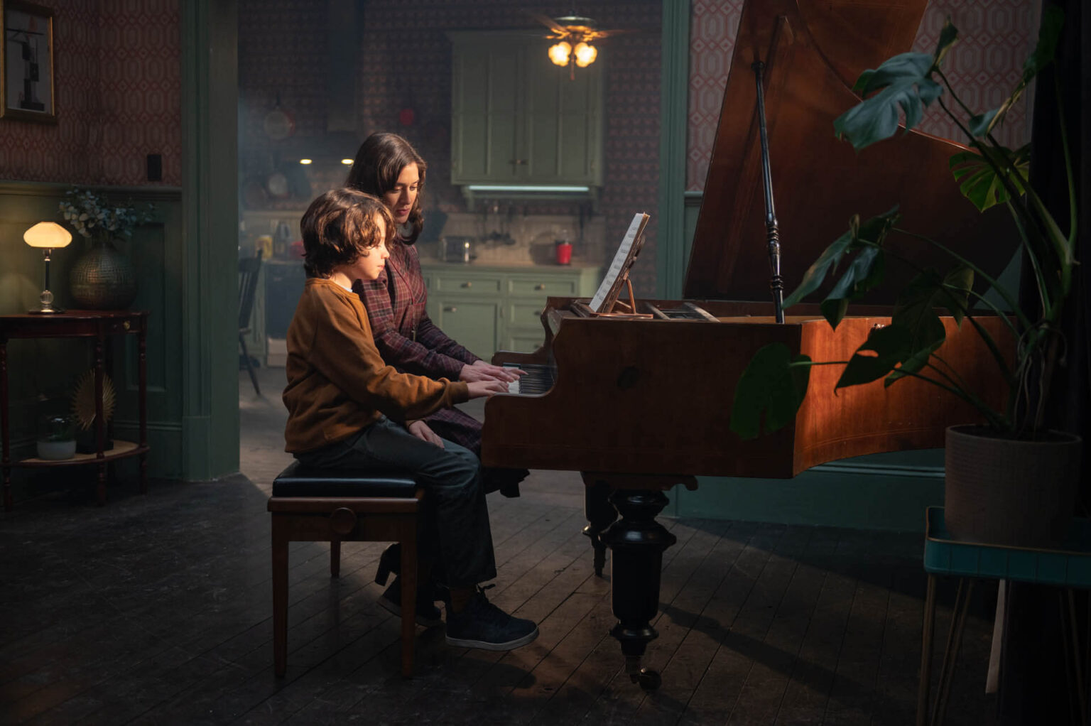 Kobieta i chłopiec siedzą razem przy fortepianie w pokoju w stylu vintage, z ciepłym oświetleniem i zielonymi ścianami, podczas gdy kobieta uczy chłopca gry.