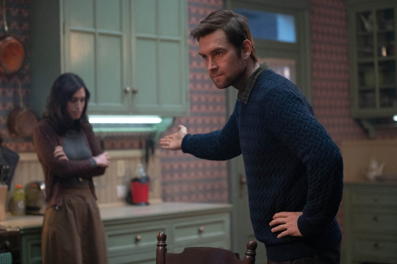 Mężczyzna w niebieskim swetrze gestykuluje ze złością w stronę kobiety ze złożonymi ramionami stojącej przy kuchennym blacie. Kobieta patrzy w dół, unikając kontaktu wzrokowego. Scenerią jest kuchnia w stylu vintage z zielonymi szafkami.