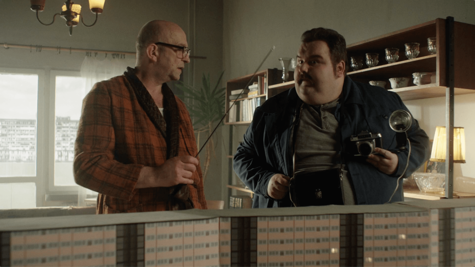 Dwóch mężczyzn stoi w pomieszczeniu; jeden w okularach i kraciastej szacie trzyma wskaźnik, a drugi w niebieskiej kurtce trzyma zabytkowy aparat. Stoją naprzeciwko siebie obok półki i modelu budynku.