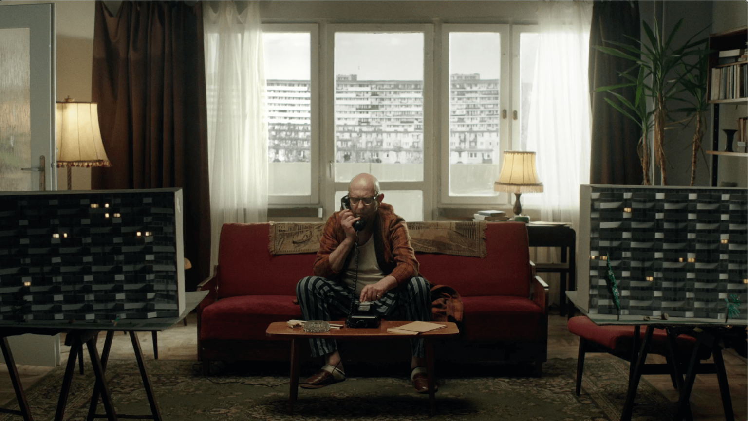 Mężczyzna siedzi na czerwonej sofie w słabo oświetlonym salonie i rozmawia przez telefon. Stoi naprzeciwko dwóch tablic z wzorami, otoczony zabytkowymi lampami, roślinami doniczkowymi, regałami na książki i dużymi oknami, na których widać bloki mieszkalne.