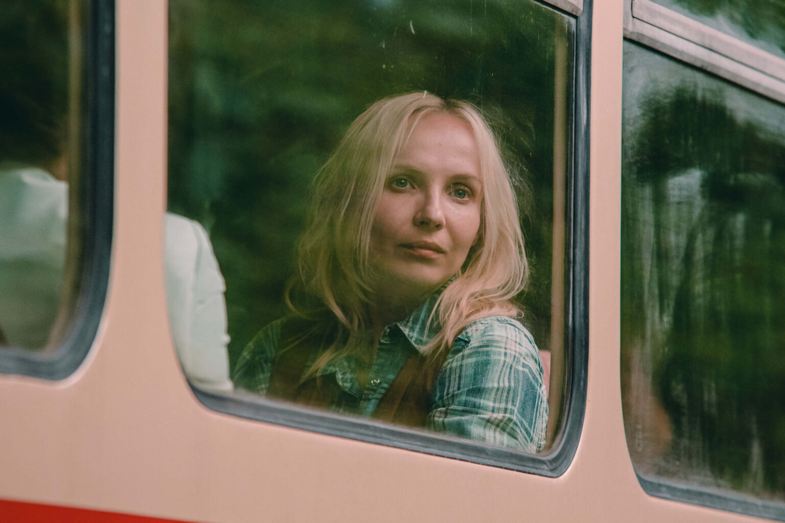Kobieta o blond włosach i kraciastej koszuli wygląda przez okno autobusu, jej twarz delikatnie odbija się w szybie. Scena na zewnątrz jest rozmazana z zielonymi tonami, co sugeruje naturalne otoczenie.