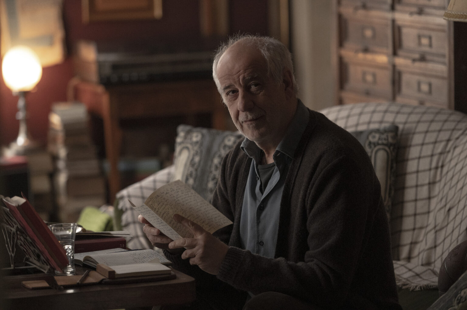 Starszy mężczyzna z siwymi włosami i brodą siedzi na sofie w przytulnym, słabo oświetlonym pokoju, trzyma otwartą książkę i patrzy w stronę kamery. Na stole przed nim stoi szklanka wody i więcej książek.