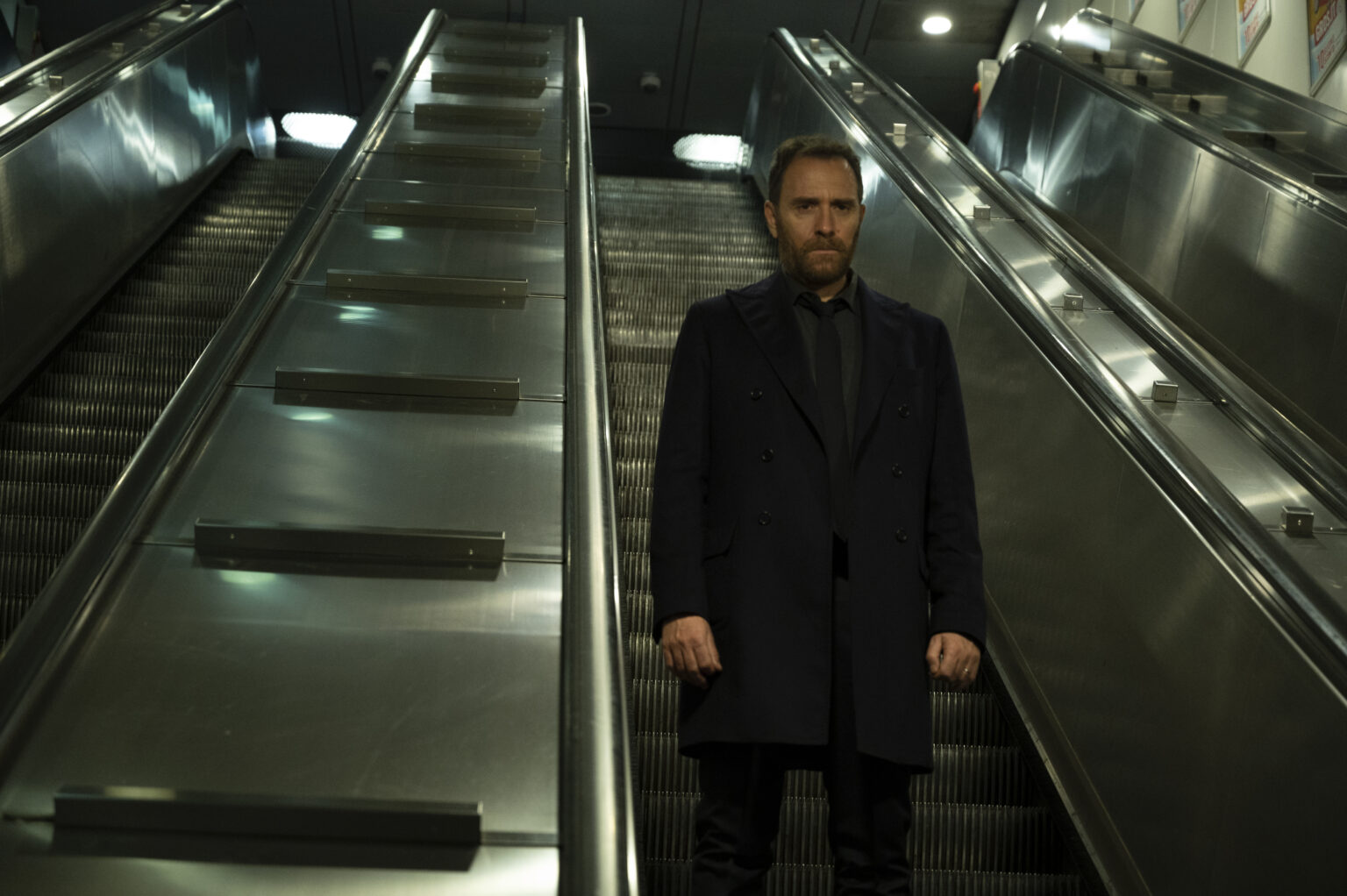 Mężczyzna w ciemnym płaszczu stoi sam na dole metalowych schodów ruchomych w słabo oświetlonym pomieszczeniu, po obu stronach którego znajdują się puste schody ruchome.