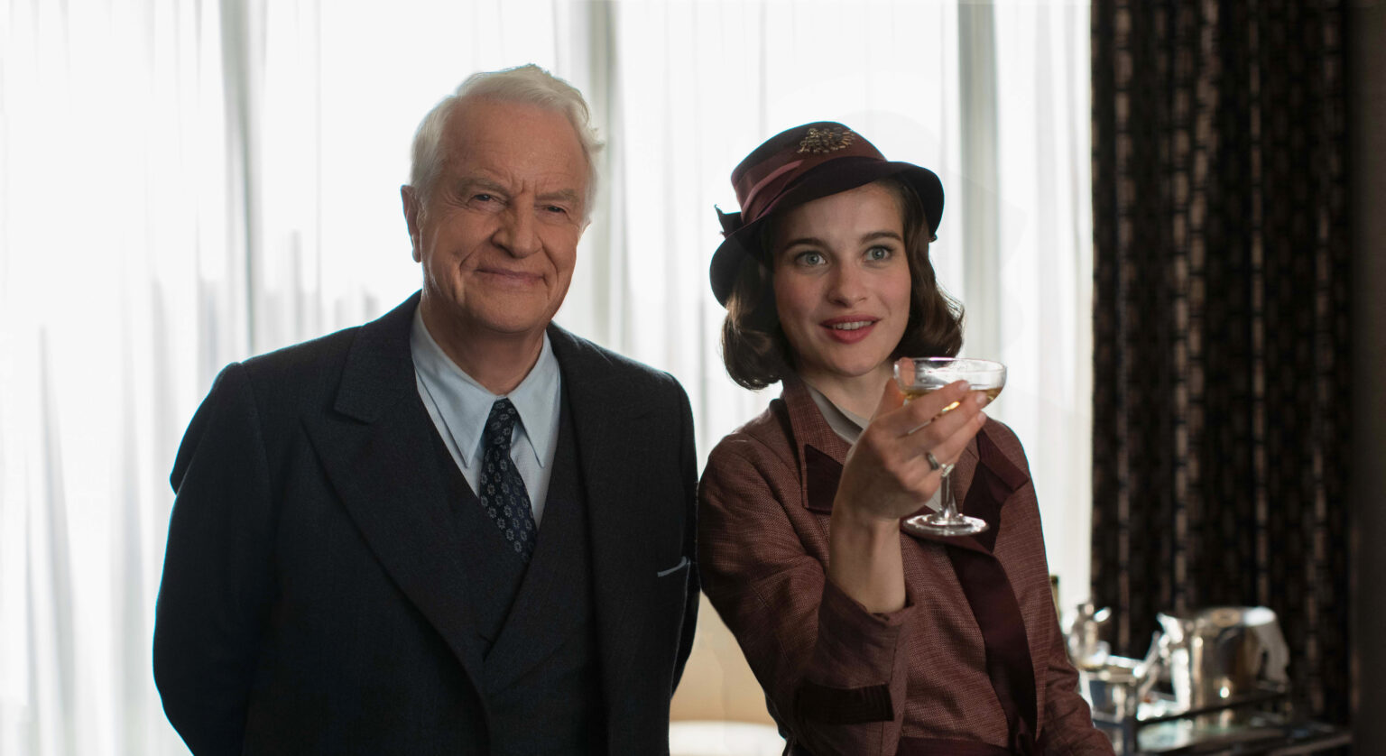 Starszy mężczyzna w garniturze stoi obok uśmiechniętej kobiety w stroju vintage i kapeluszu, która trzyma drinka w toaście. Są w środku, a światło wpada przez cienkie firanki za nimi.