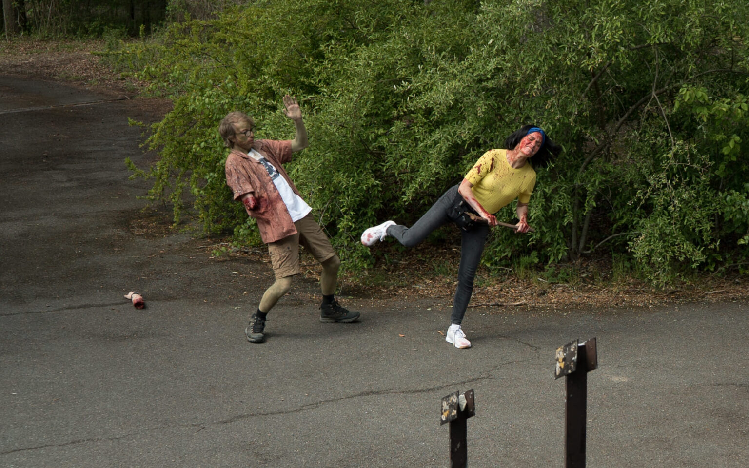 Dwie osoby w kostiumach zombie odgrywają dramatyczną scenę na wybrukowanej ścieżce otoczonej zielenią. Jedna osoba zatacza się do tyłu z uniesionymi rękami, podczas gdy druga, pokryta sztuczną krwią, kopie jedną nogą.