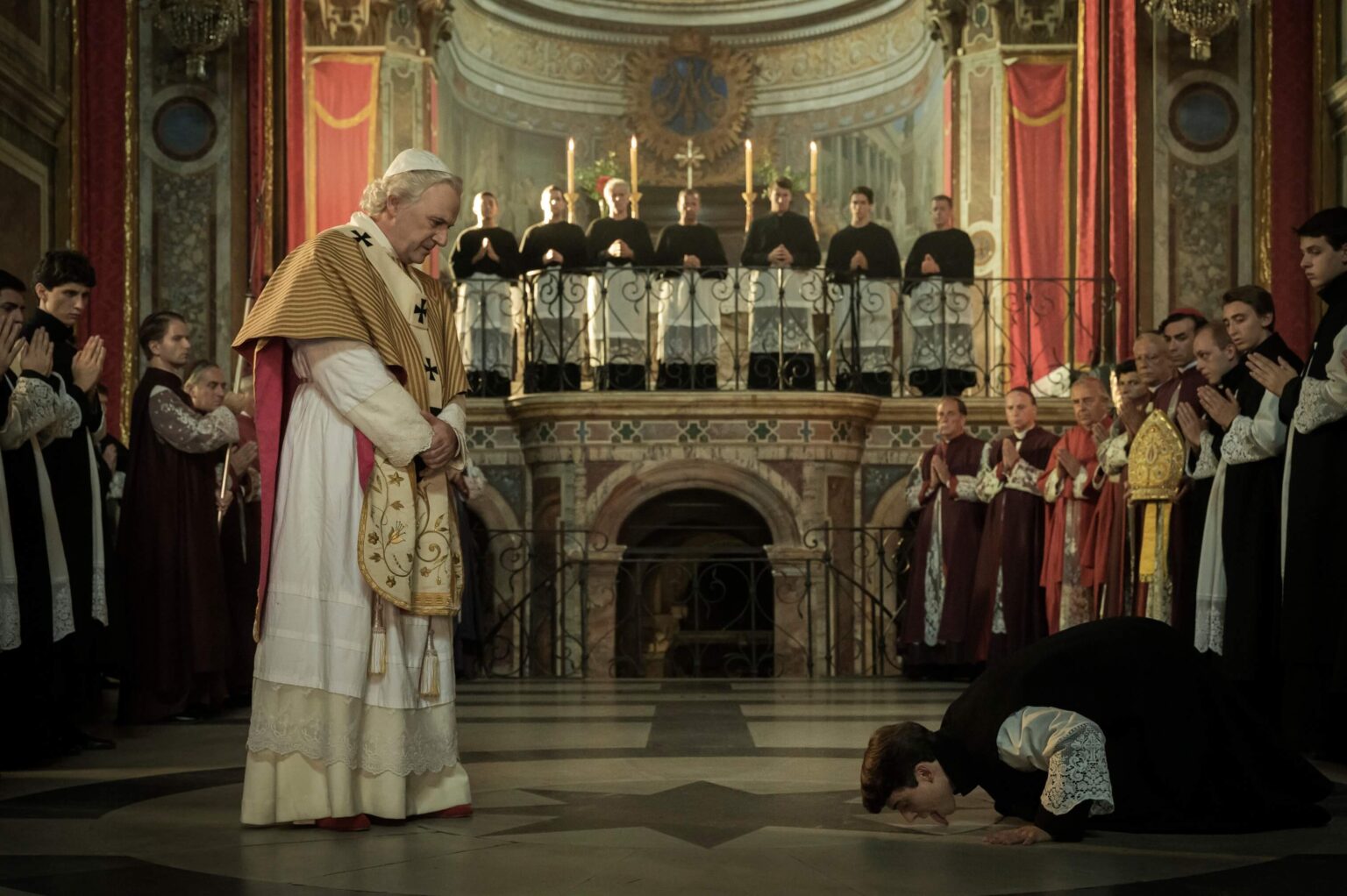 Osoba ubrana w strój religijny klęka i kłania się przed wysokim rangą duchownym w ozdobnych szatach, otoczona duchownymi i gapiami w wielkim, oświetlonym świecami kościele z czerwonymi zasłonami i misternymi dekoracjami.