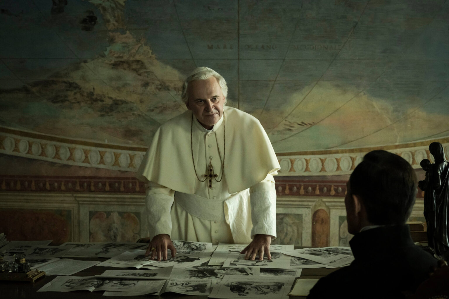 Starszy mężczyzna w białej papieskiej szacie stoi za stołem pokrytym papierami i patrzy na siedzącą osobę w ciemnym pokoju, na ścianie którego wisi duża mapa świata.