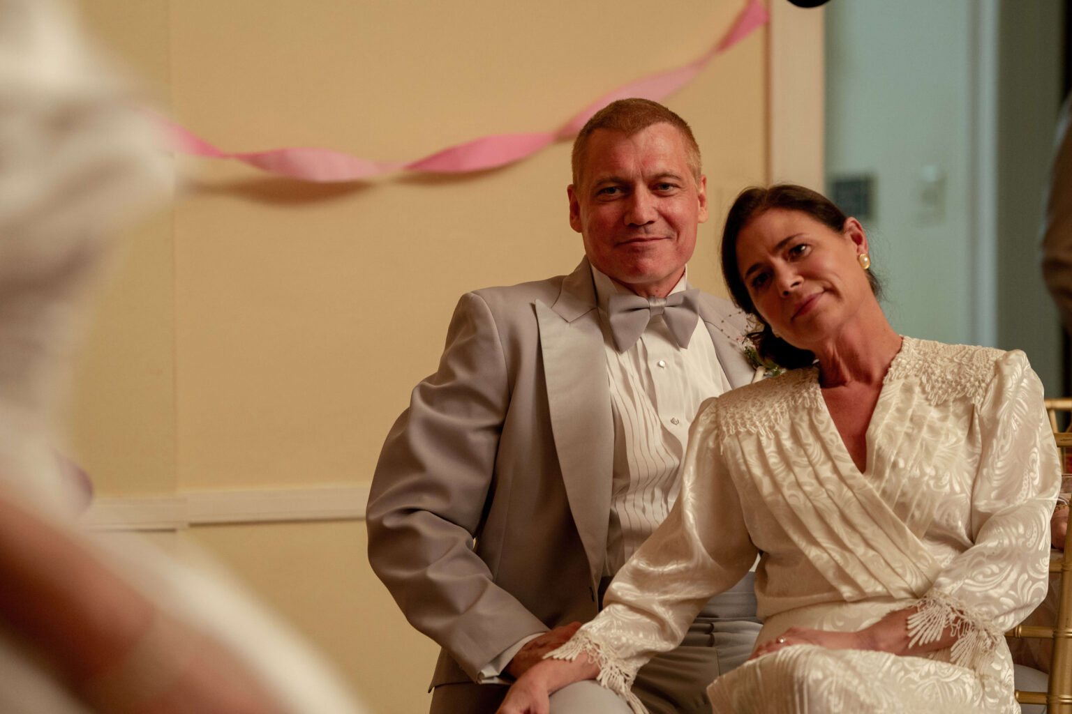 Mężczyzna w jasnoszarym smokingu i kobieta w białej sukience w stylu vintage siedzą blisko siebie, uśmiechając się ciepło. Różowe serpentyny zdobią tło, sugerując wydarzenie o charakterze celebrującym.