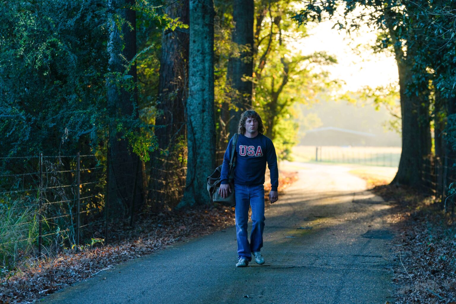 Osoba z długimi włosami ubrana w bluzę USA idzie samotnie po wiejskiej drodze obsadzonej drzewami o wschodzie słońca, niosąc torbę. Światło słoneczne przesącza się przez drzewa, tworząc spokojną, ciepłą atmosferę.
