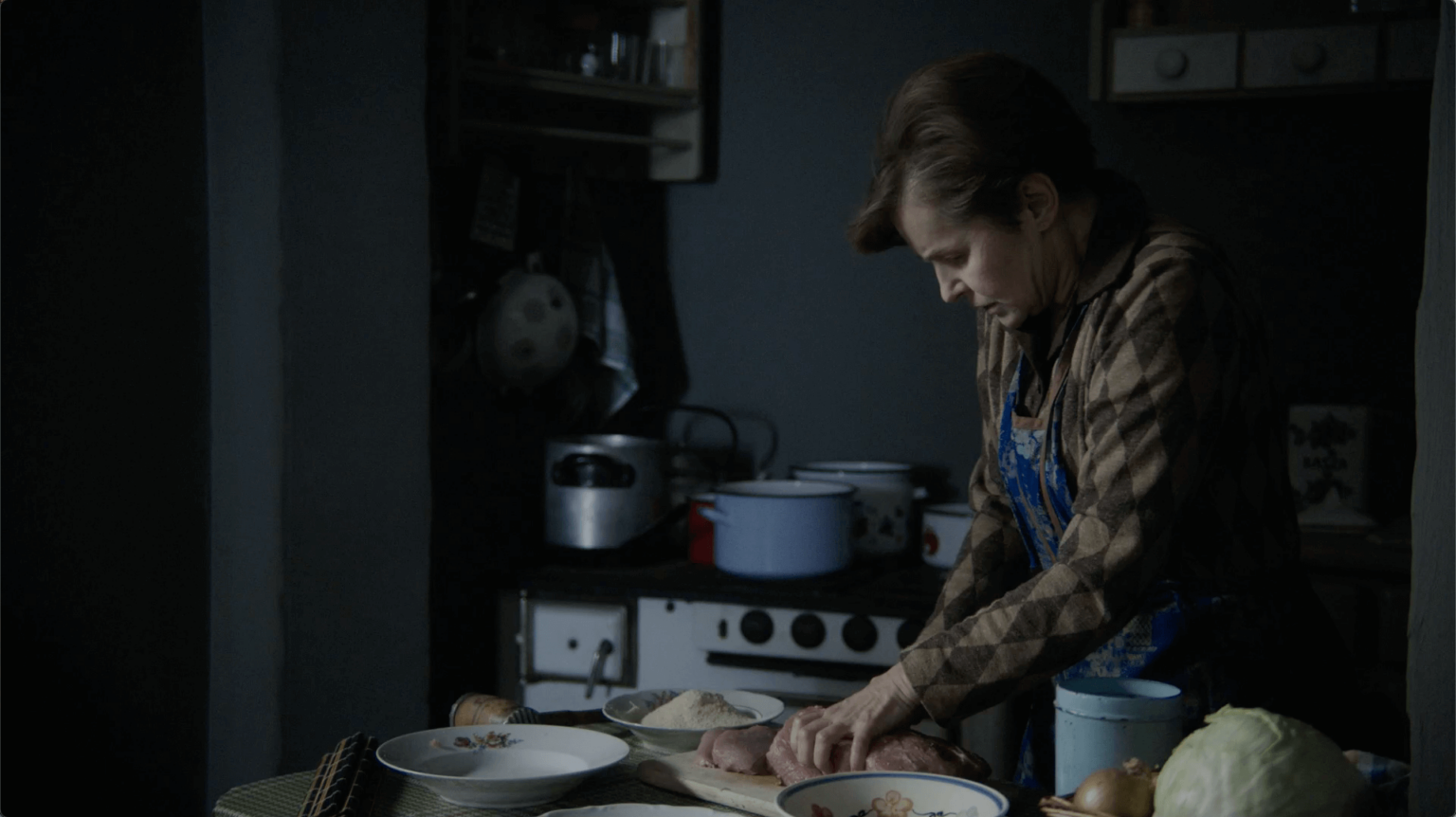 Kobieta ubrana w fartuch przygotowuje jedzenie na kuchennym blacie, otoczona miskami i warzywami, z kuchenką i garnkami na tle słabo oświetlonego tła.