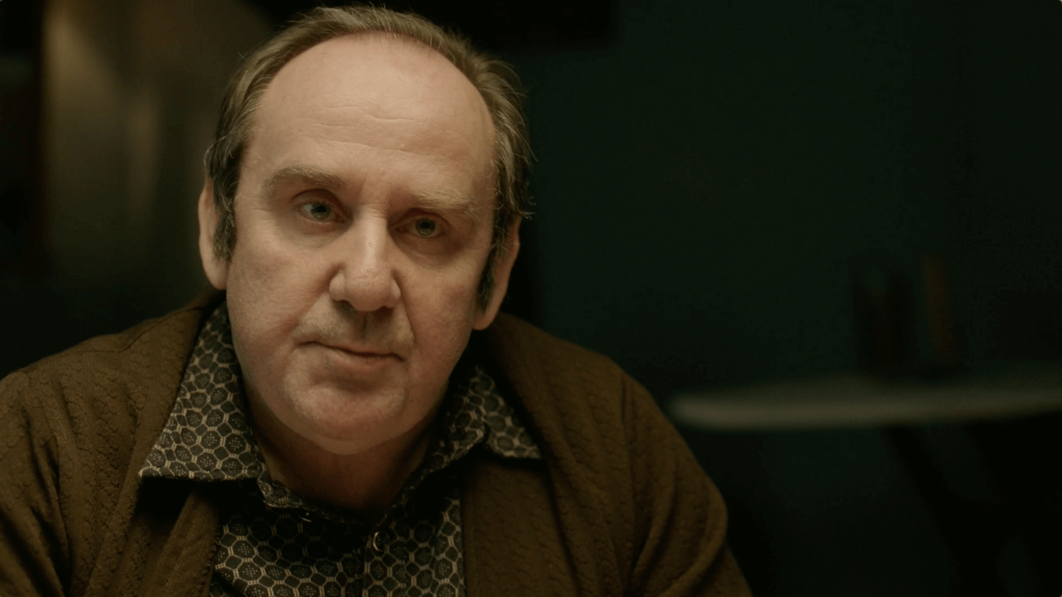 Mężczyzna w średnim wieku z przerzedzonymi włosami i wzorzystą koszulą siedzi przy stole, wyglądając poważnie. Tło jest ciemne i nieostre.