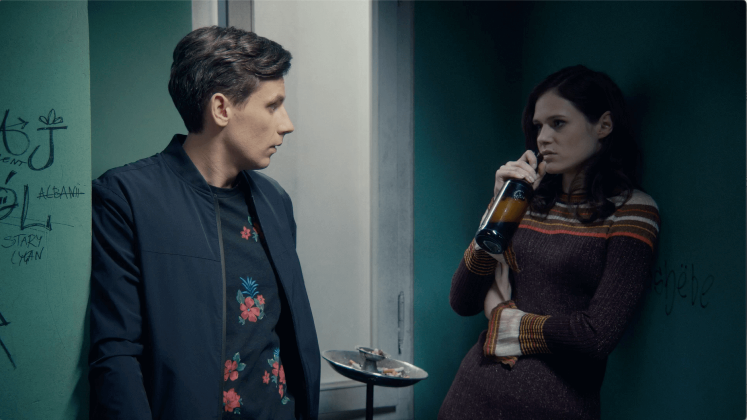 Mężczyzna w ciemnej kurtce staje naprzeciwko kobiety w pasiastym swetrze, która opiera się o ścianę i pije z butelki. Znajdują się w słabo oświetlonym pomieszczeniu z graffiti na zielonych ścianach i małą tacą na stojaku w pobliżu.