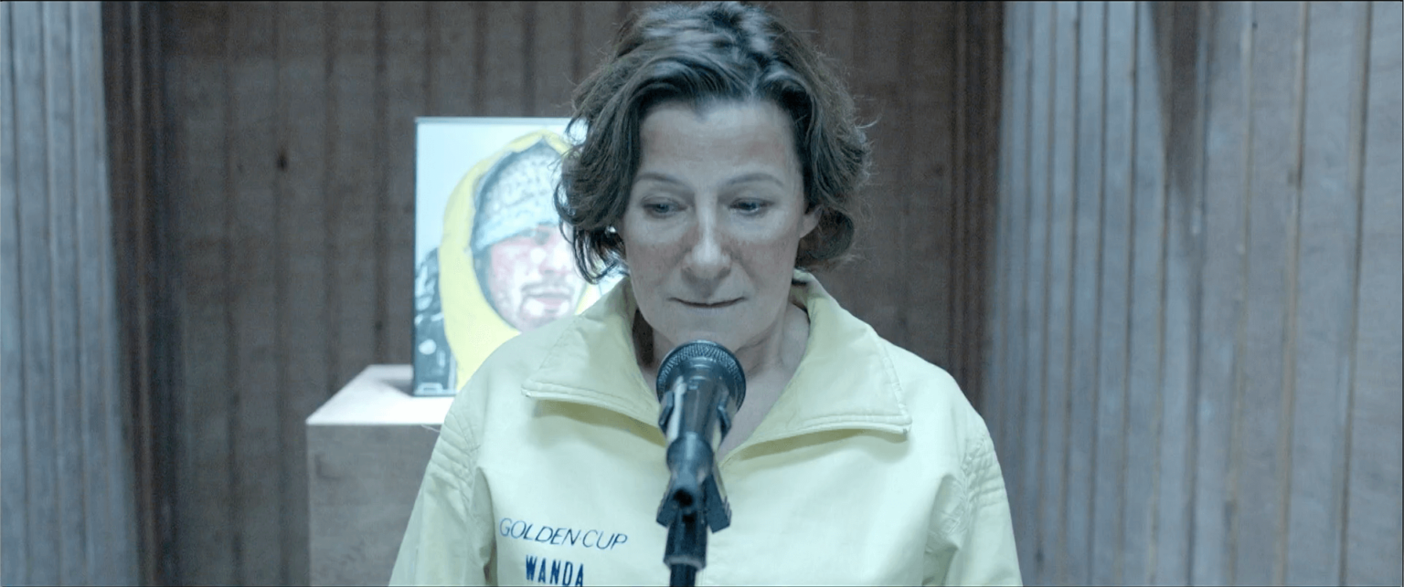 Kobieta z krótkimi brązowymi włosami, ubrana w jasnożółtą kurtkę, stoi przed mikrofonem. Za nią znajduje się oprawiona praca artystyczna przedstawiająca twarz i żółte tło, wystawiona na drewnianym cokole.