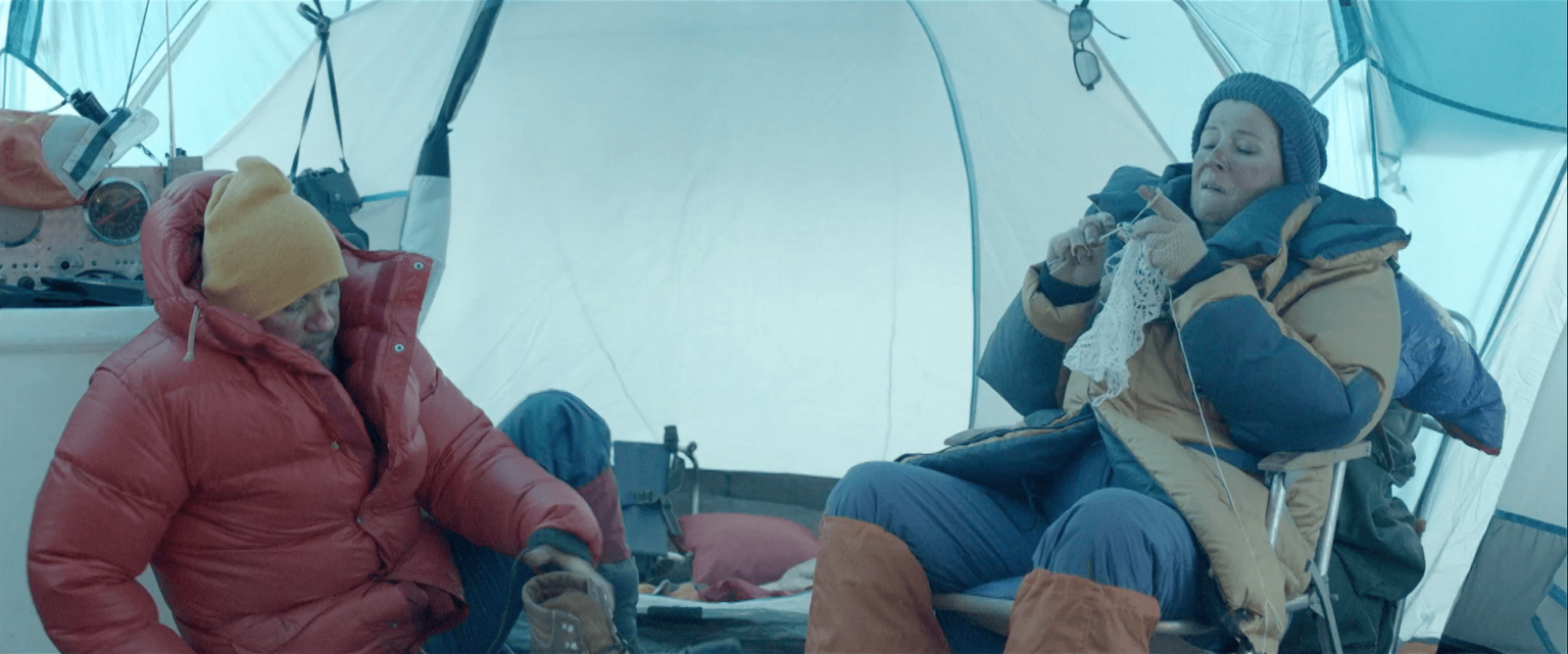 Dwie osoby w zimowych kurtkach siedzą w namiocie; jedna robi na drutach, a druga relaksuje się z kubkiem w otoczeniu odzieży na zimną pogodę i sprzętu kempingowego.