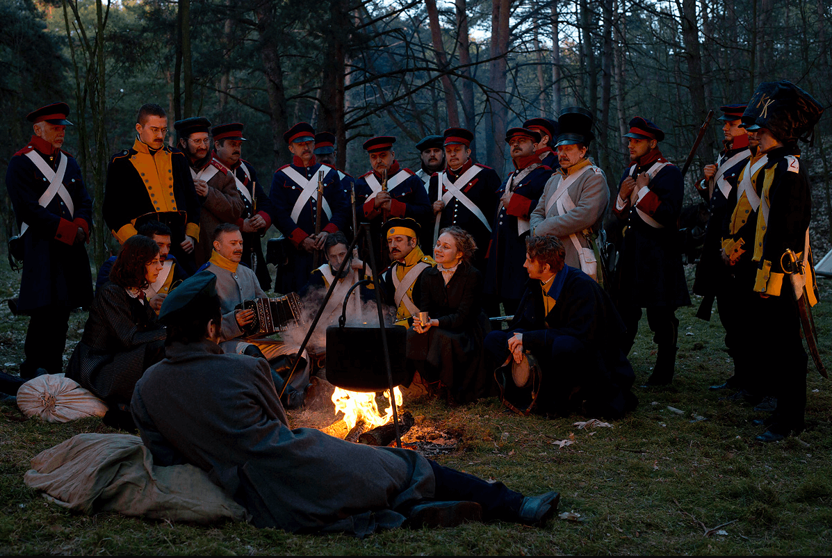 Grupa ludzi w historycznych mundurach wojskowych i ubraniach z epoki zbiera się wokół ogniska w lesie. Część osób siedzi, część stoi, a nad płomieniami wisi czajnik.