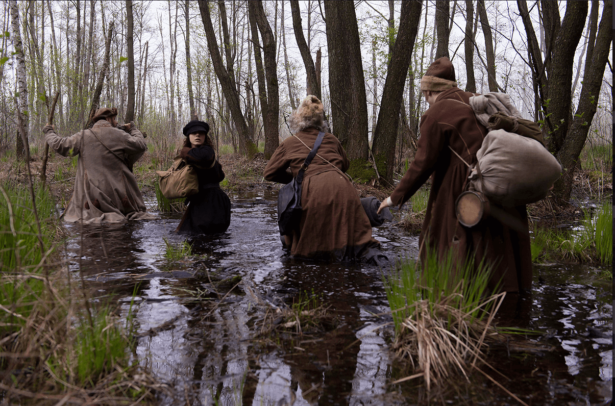 Cztery osoby ubrane w grube brązowe płaszcze brną przez błotnisty, zalany wodą las, niosąc torby i dobytek, otoczone wysokimi trawami i bezlistnymi drzewami.