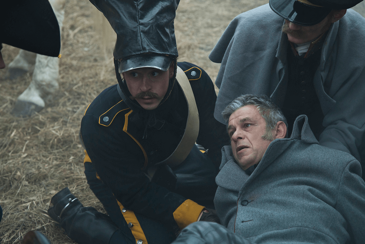 Żołnierz w ciemnym mundurze klęczy obok starszego mężczyzny w szarym płaszczu leżącego na ziemi, podczas gdy inny mężczyzna w mundurze pochyla się nad nimi. Scena wydaje się napięta i historyczna.