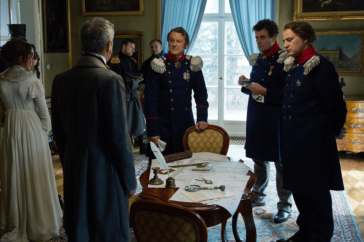 Grupa osób ubranych w XIX-wieczne mundury wojskowe i formalne stroje stoi wokół stołu pokrytego papierami i przyborami do pisania w eleganckim, słonecznym pokoju z dużymi oknami i ozdobnymi meblami.