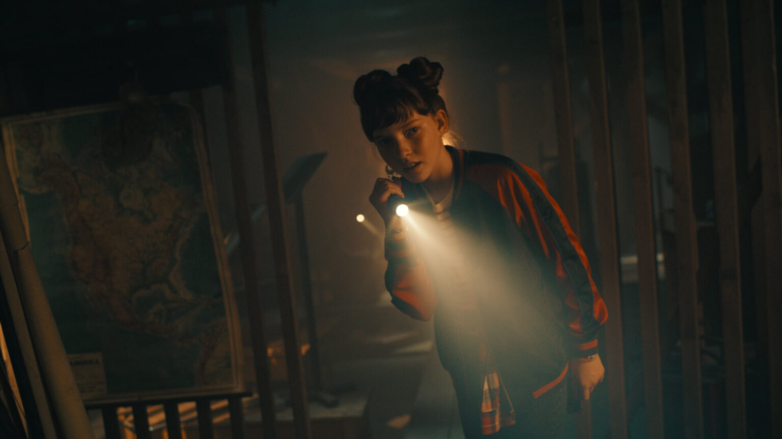 Młoda osoba z dwoma kokami we włosach stoi w ciemnym, zagraconym pokoju, świecąc latarką. W pobliżu wisi duża mapa Ameryki Północnej. Atmosfera jest tajemnicza i pełna napięcia.