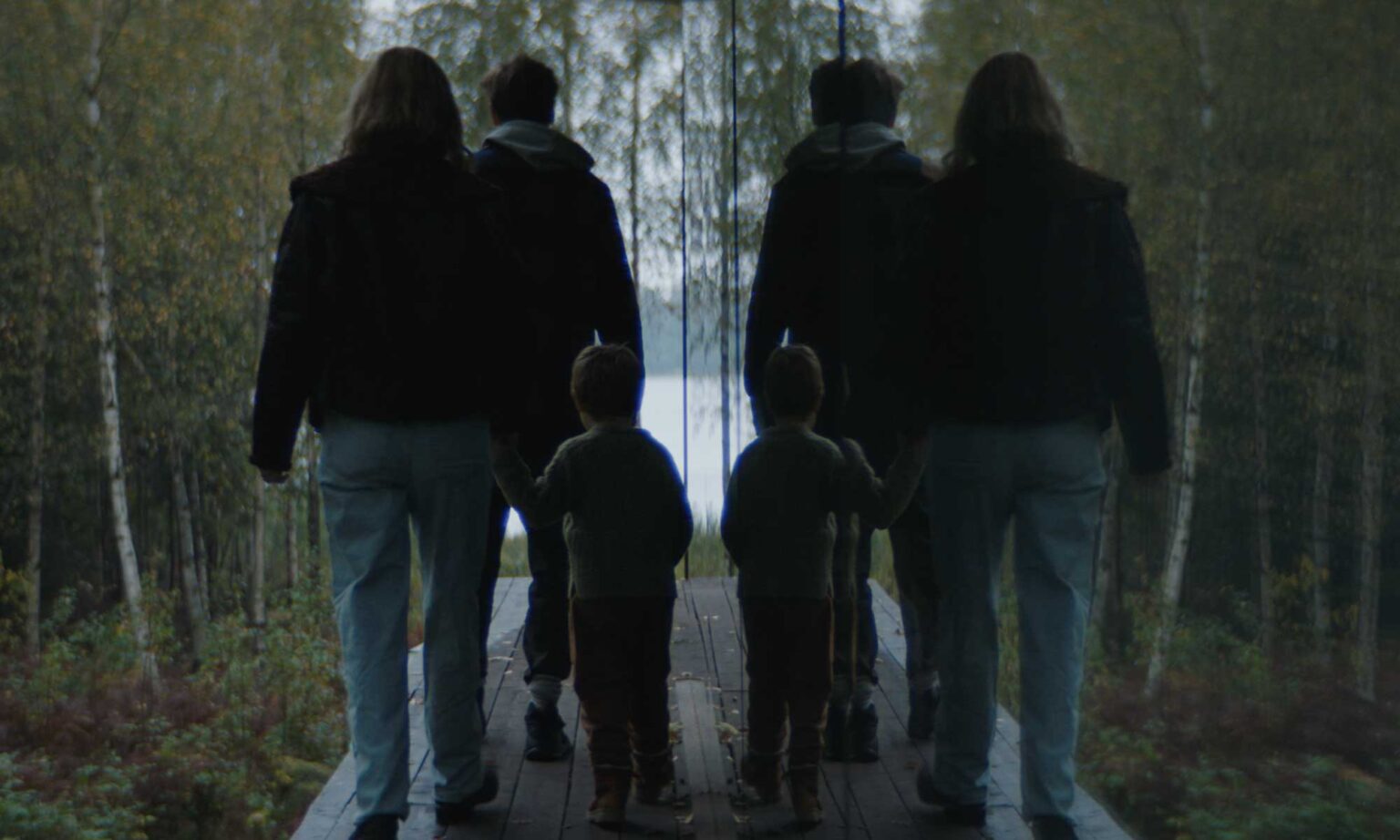 Dwoje dorosłych i dziecko idą ręka w rękę po drewnianej ścieżce przez las, ich odbicia widać w dużym lustrze po ich prawej stronie. W tle widać drzewa i fragment jeziora.