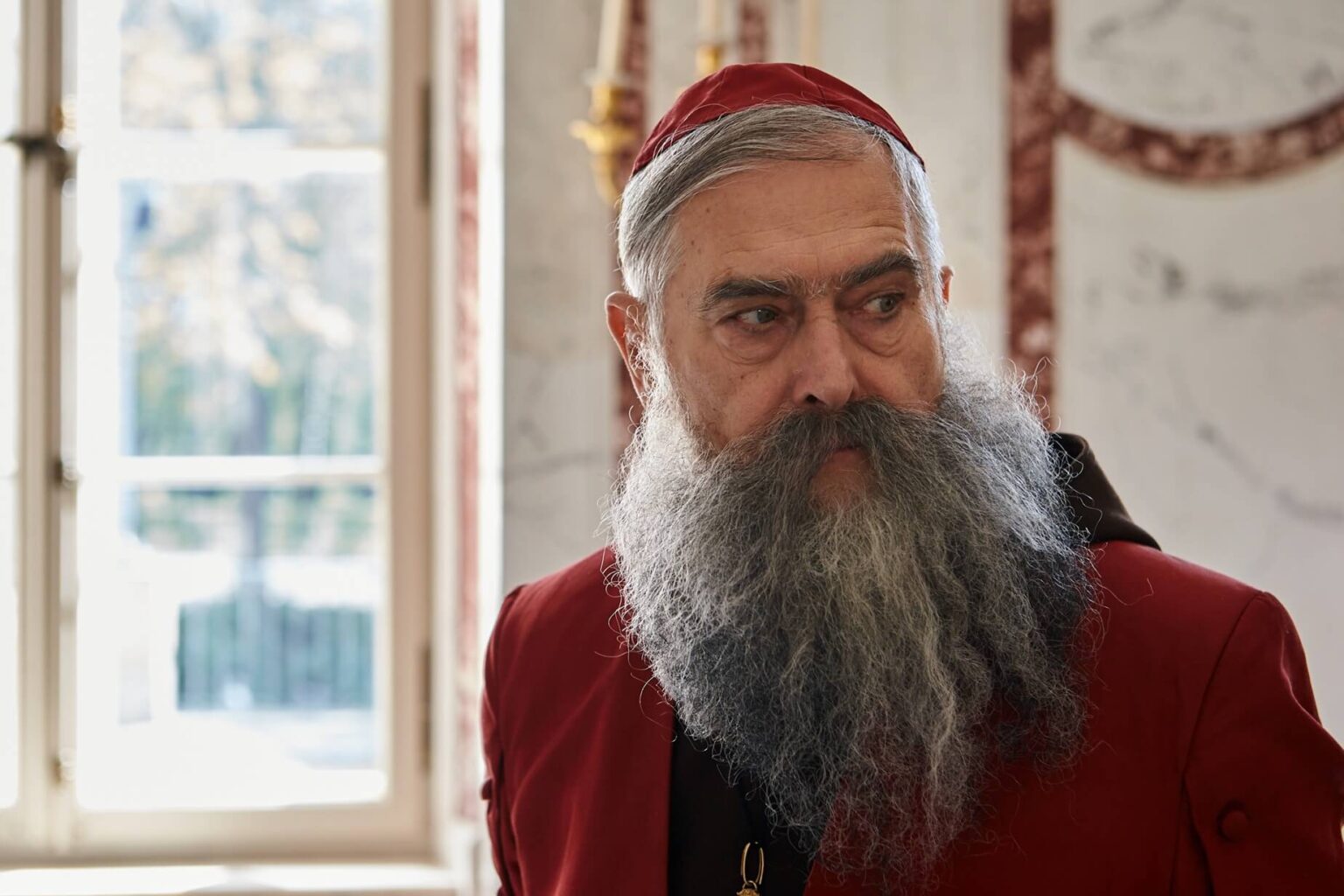 Starszy mężczyzna z długą, siwą brodą i wąsami nosi czerwoną szatę i czapkę, stoi w pomieszczeniu przy oknie, przez które wpada światło. W tle widoczne są ściany z marmurowym wzorem i elegancki wystrój.