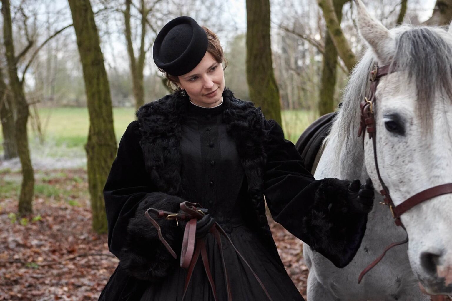Kobieta w stroju z epoki wiktoriańskiej i czarnym kapeluszu delikatnie trzyma lejce białego konia w zalesionym terenie z nagimi drzewami i opadłymi liśćmi na ziemi.