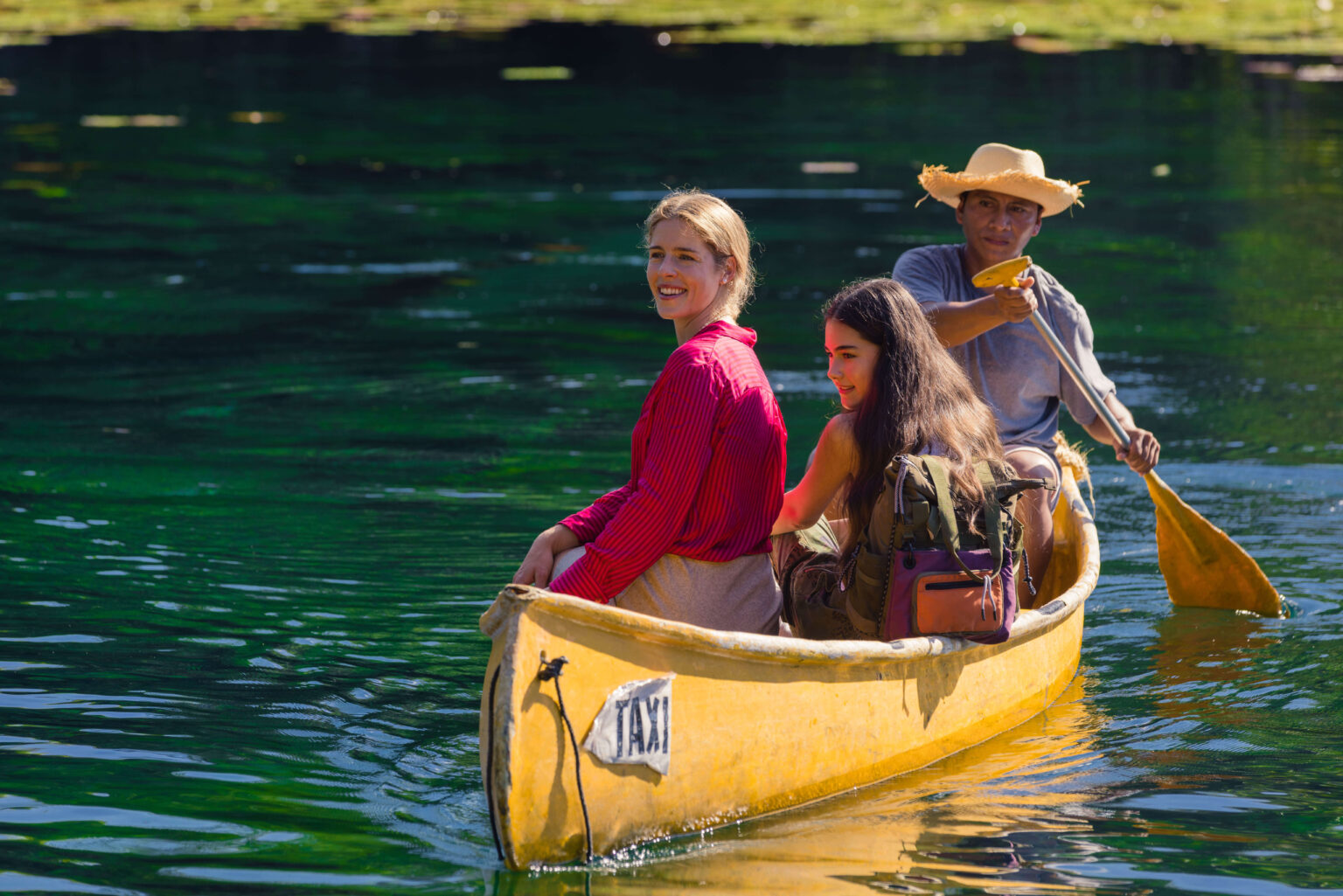 Trzy osoby płyną żółtą łodzią po spokojnym, zielonkawym jeziorze. Dwie kobiety siedzą z przodu, jedna uśmiecha się do kamery, podczas gdy mężczyzna w słomkowym kapeluszu wiosłuje z tyłu. W tle widać drzewa i rośliny wodne.