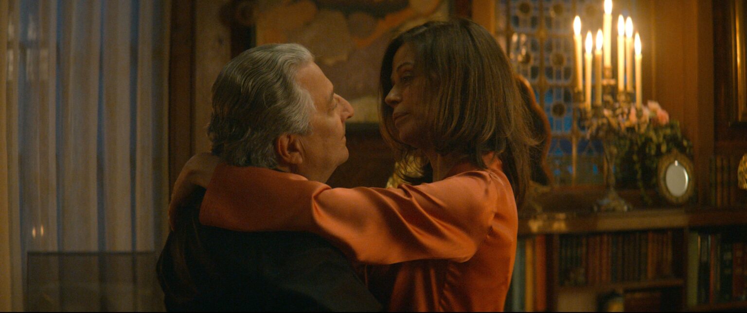 Mężczyzna i kobieta obejmują się blisko w ciepło oświetlonym pokoju z regałami na książki, świecami i ozdobnym szkłem w tle. Kobieta ma na sobie pomarańczową bluzkę i patrzy na mężczyznę, który ma siwiejące włosy i nosi ciemną kurtkę.