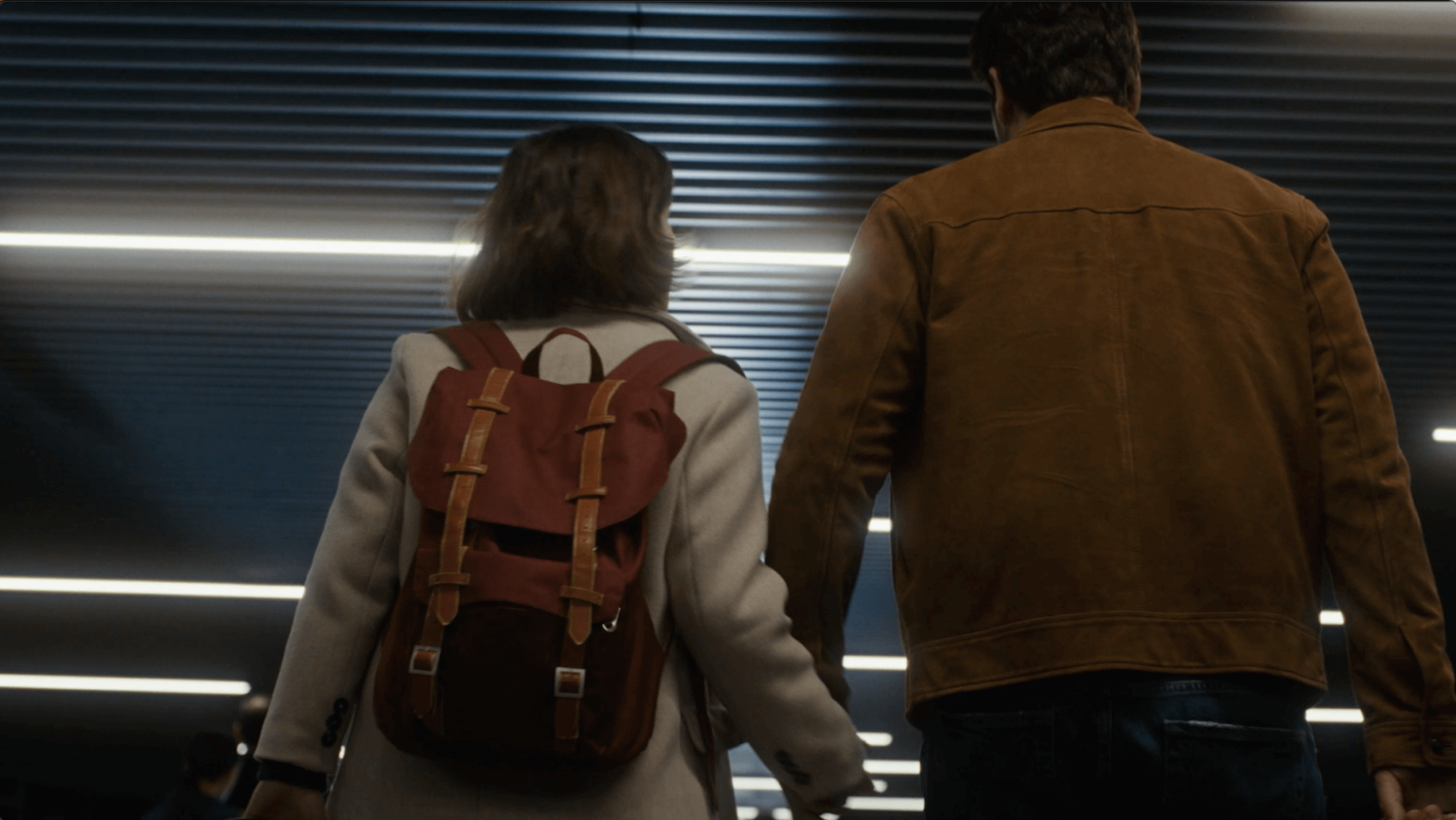 Kobieta z czerwonym plecakiem i mężczyzna w brązowej kurtce trzymają się za ręce, idąc w pomieszczeniu pod jasnymi, poziomymi światłami sufitowymi. Zdjęcie zostało zrobione od tyłu.