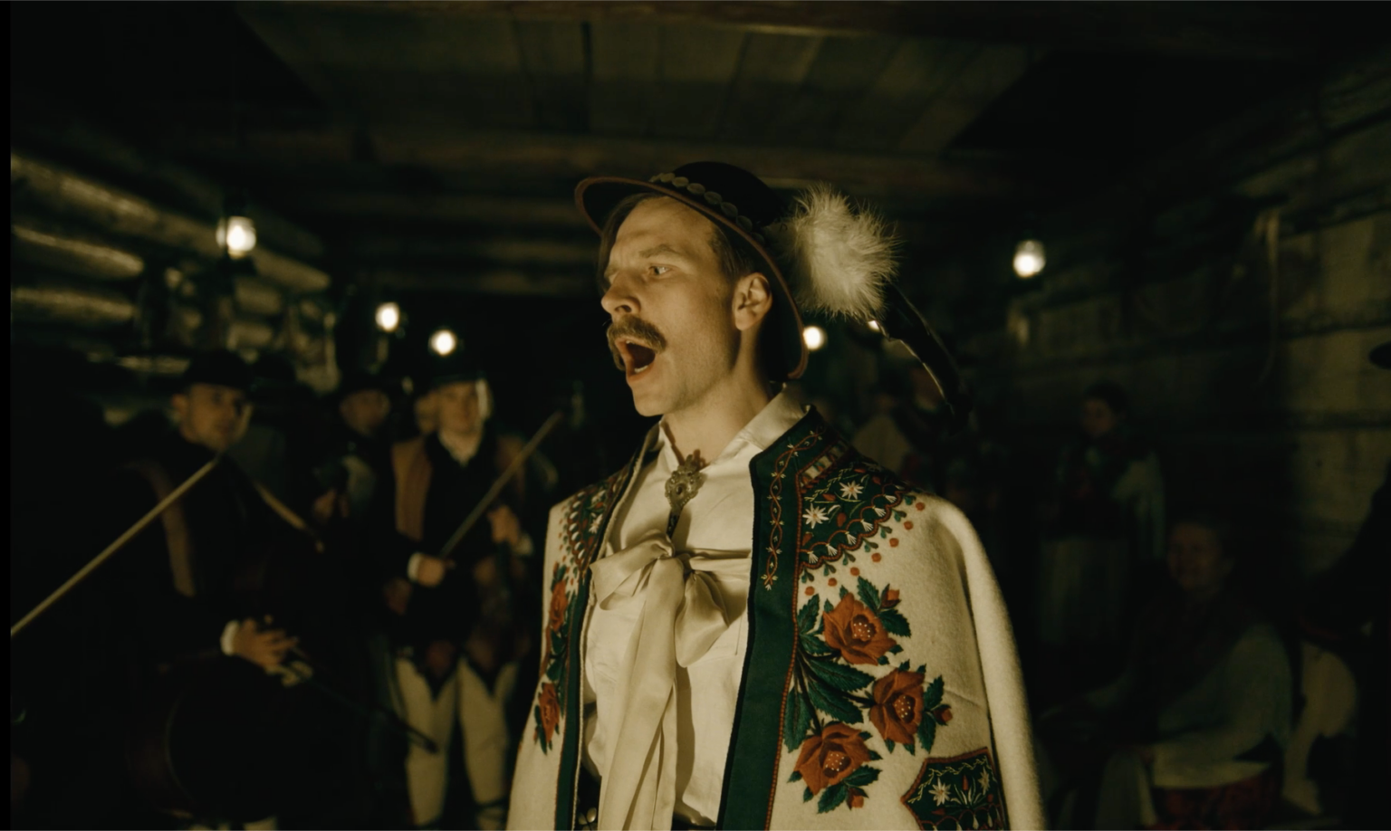Mężczyzna ubrany w tradycyjny, haftowany strój ludowy z kwiatowymi wzorami i czapkę z piórami śpiewa namiętnie w słabo oświetlonym drewnianym pokoju, podczas gdy inni, ubrani w podobne stroje, grają na instrumentach w tle.