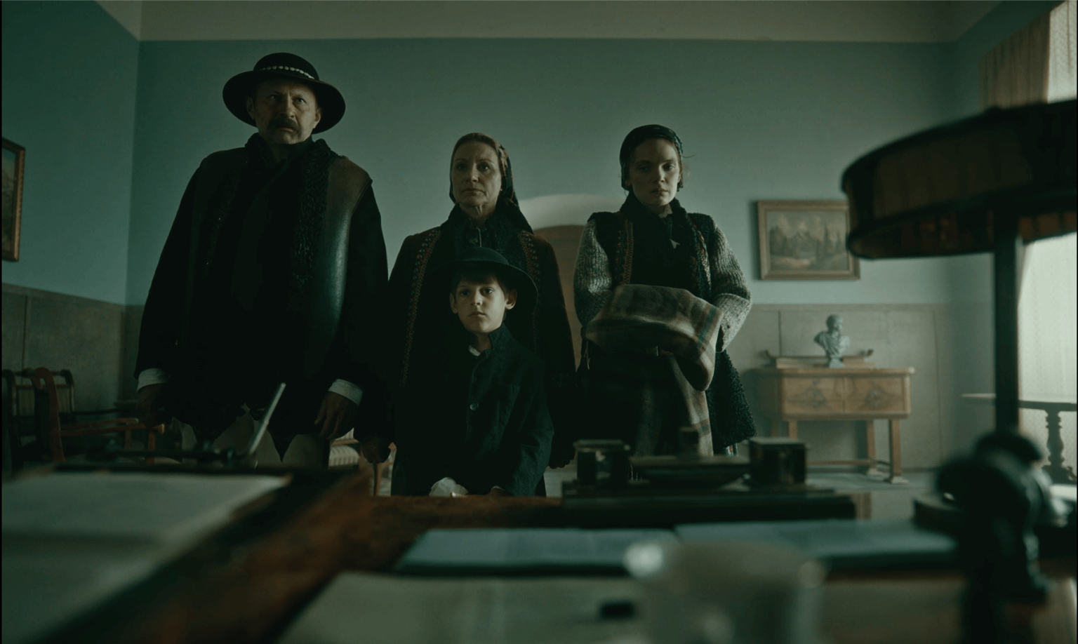 Cztery osoby, trzy osoby dorosłe i jedno dziecko, ubrani w staromodne, ciemne ubrania, stoją uroczyście w słabo oświetlonym pokoju, patrząc w stronę biurka na pierwszym planie. Sceneria wydaje się historyczna i poważna.