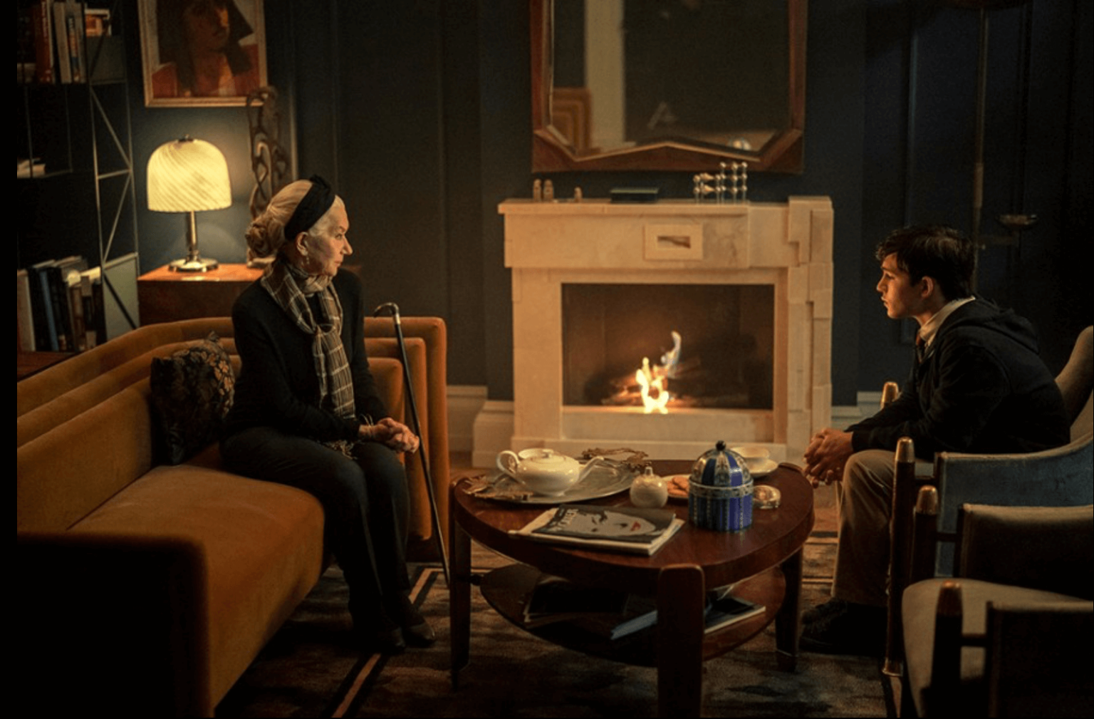 Starsza kobieta i młody mężczyzna siedzą naprzeciwko siebie w przytulnym salonie z rozpalonym kominkiem, ciepłym oświetleniem i stolikiem kawowym zastawionym herbatą i przekąskami między nimi.