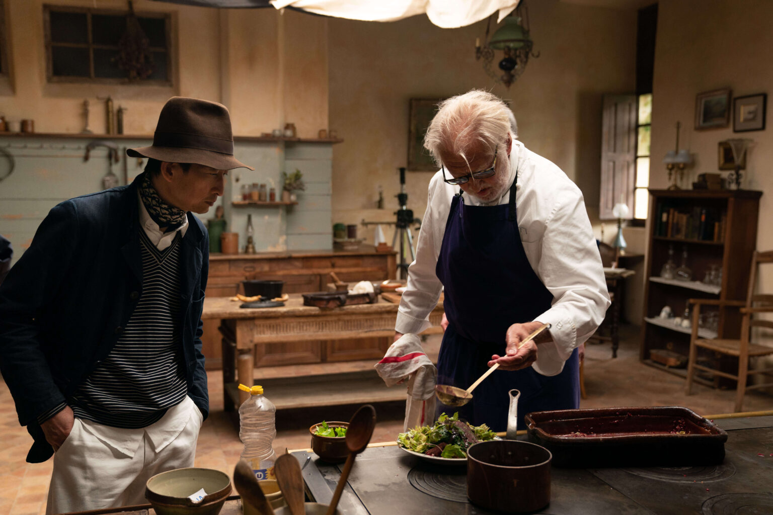 Starszy mężczyzna w fartuchu podaje jedzenie na talerzu w rustykalnej kuchni, podczas gdy młodszy mężczyzna w czapce i szaliku uważnie się temu przygląda. Kuchnia jest wypełniona drewnianymi meblami i zabytkowymi dekoracjami.