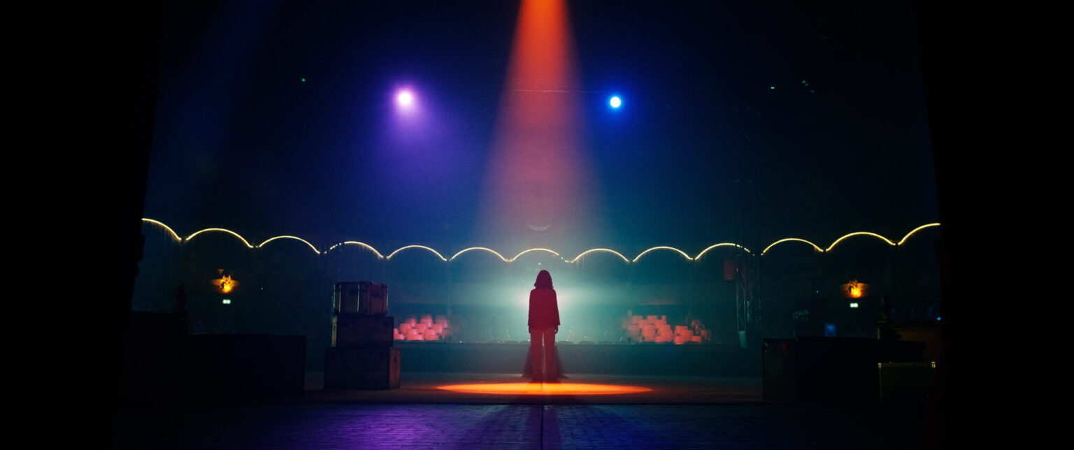 Samotna osoba stoi pod jasnopomarańczowym reflektorem pośrodku ciemnego, pustego miejsca z kolorowymi światłami i świecącymi łukami w tle.