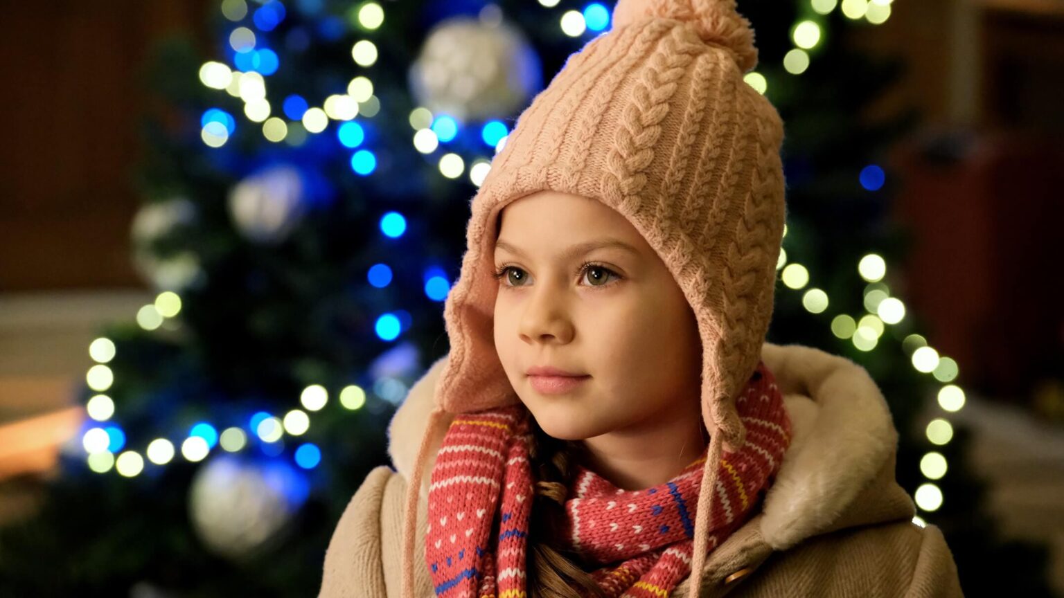 Małe dziecko w ciepłym płaszczu, różowej czapce i kolorowym szaliku stoi przed udekorowaną choinką z niebieskimi i białymi światełkami, patrząc w bok ze spokojnym wyrazem twarzy.