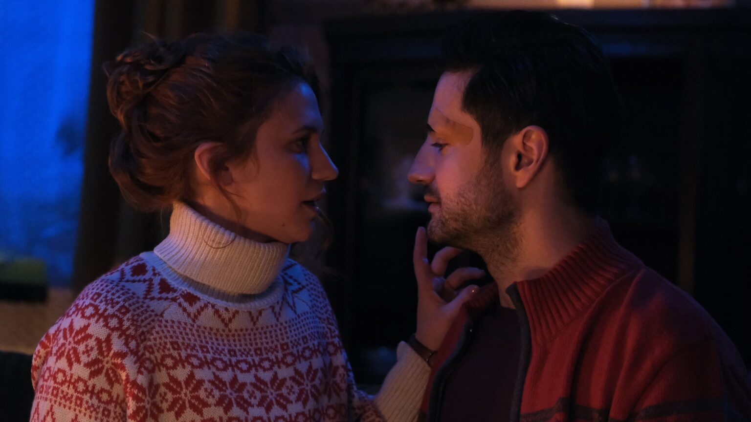 Kobieta w biało-czerwonym wzorzystym swetrze delikatnie dotyka twarzy mężczyzny, gdy patrzą sobie w oczy w słabo oświetlonym, przytulnym pokoju. Atmosfera wydaje się intymna i ciepła.